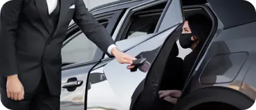 A driver in a black suit opens a black car door for a female passenger wearing a black face mask.