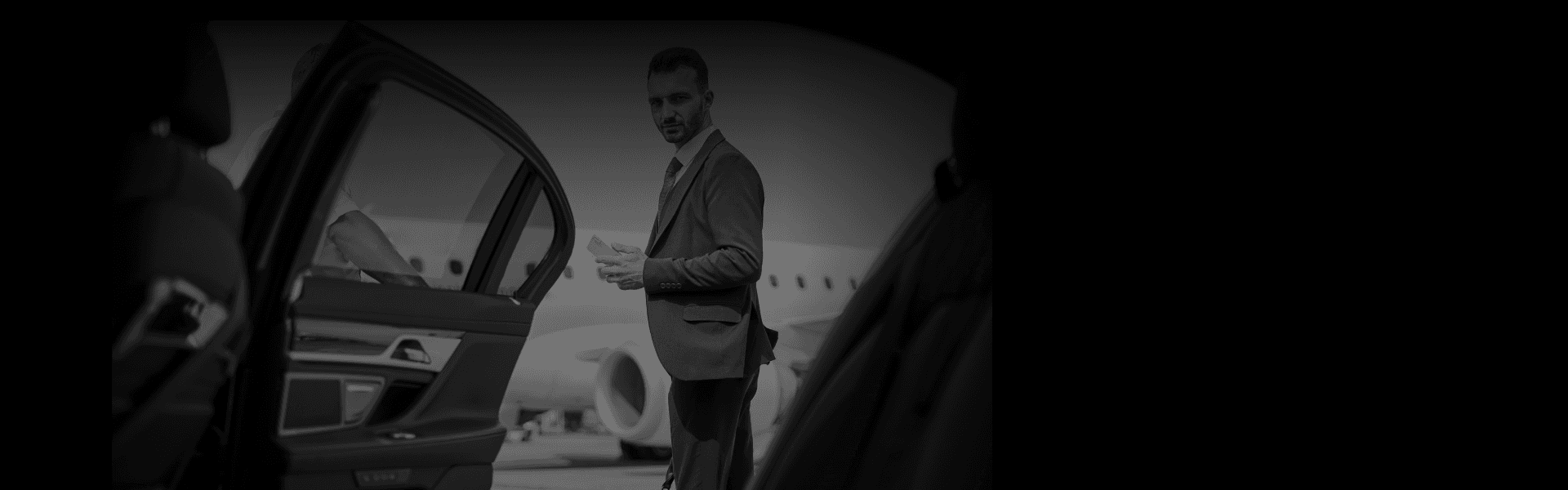 Business traveler in a grey suit exits a luxury black car on the airport tarmac, highlighting premium airport car service from a side rear-door angle.