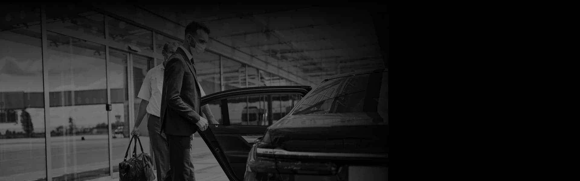 Two men in suits, one masked, load luggage beside a sleek black car at curbside—angled for a seamless airport transfer experience.