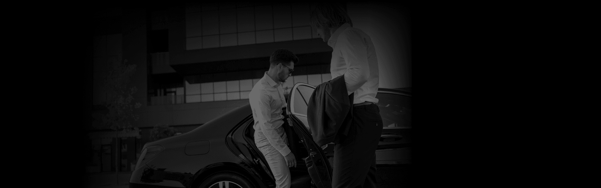 City-To-City transfer scene with two suited men beside a sleek black car, captured at a slight rear side angle near a modern office facade.