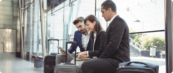 Three professionals with luggage work on a laptop and tablet while seated in a bright airport terminal lounge.