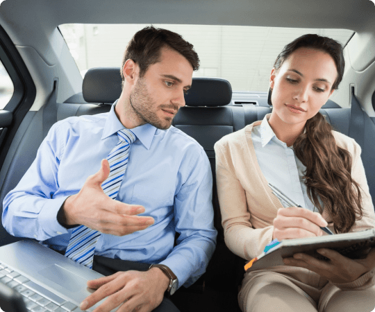 Businessman and businesswoman collaborate with a laptop and notebook in the backseat of a luxury car, showcasing mobile productivity.