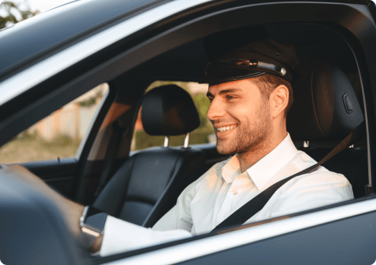 Young professional chauffeur in uniform smiles confidently while seated in a premium black car, ready for a smooth ride.