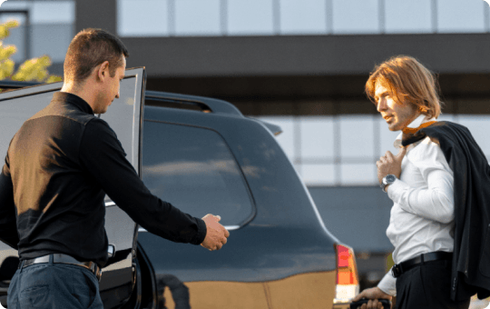 A chauffeur in black opens the SUV door for a stylish business traveler in a white shirt under warm sunlight.