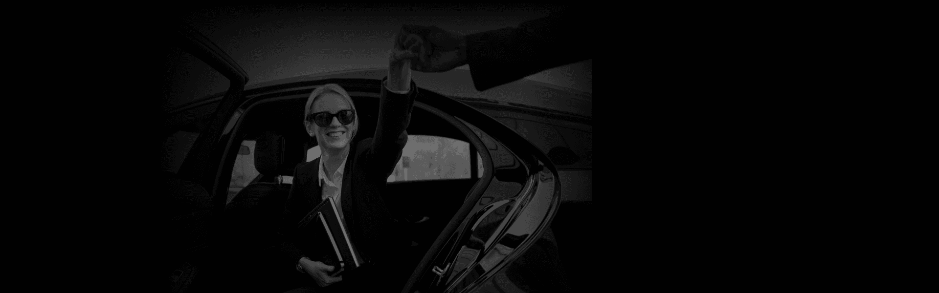 Smiling woman in sunglasses stepping out of a luxury car and reaching for a handshake, showcasing point-to-point car service with a door-side perspective.