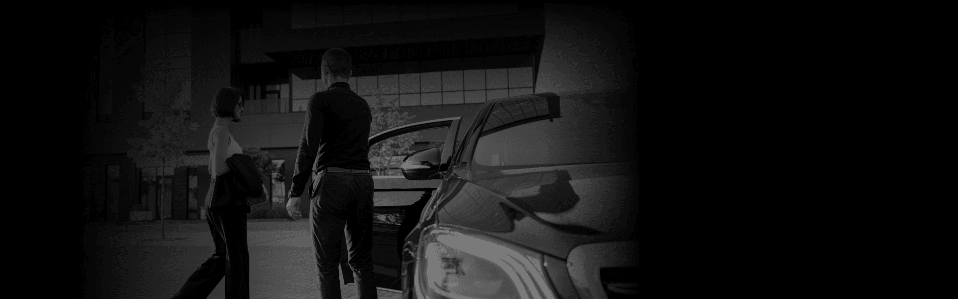 A man opens the door of a sleek black luxury car for a woman dressed elegantly, set against a modern building backdrop, ideal for Special Events.