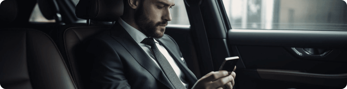 Businessman in a black suit checking phone in the rear seat of a luxury car, viewed from a side interior angle.