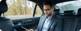 A man in formal attire is using his phone in the backseat of a luxury sedan, commuting through a scenic route.
