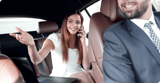 A businesswoman cheerfully gestures while talking on the phone, seated comfortably in a luxe backseat with a laptop.