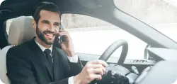 A smiling man in a suit is taking a support call while driving.