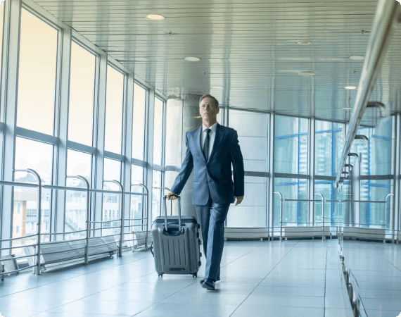 Business traveler in a navy blue suit walking confidently through a modern glass-walled airport terminal with rolling luggage.