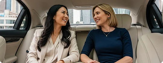 Two professional women sit in the back of a luxury car with cream-colored leather seats, smiling and looking at each other while engaged in conversation.