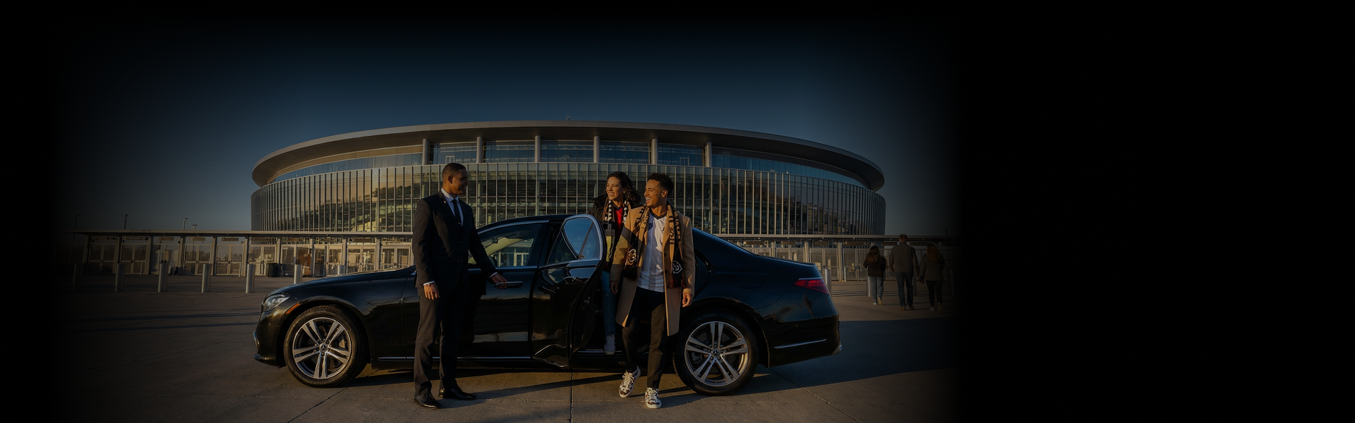 A chauffeur in a dark suit opens the door of a sleek black sedan for a smiling young couple arriving at a large, modern glass stadium under a clear sky at dusk. The couple is dressed in stylish coats and scarves, appearing excited for an event.