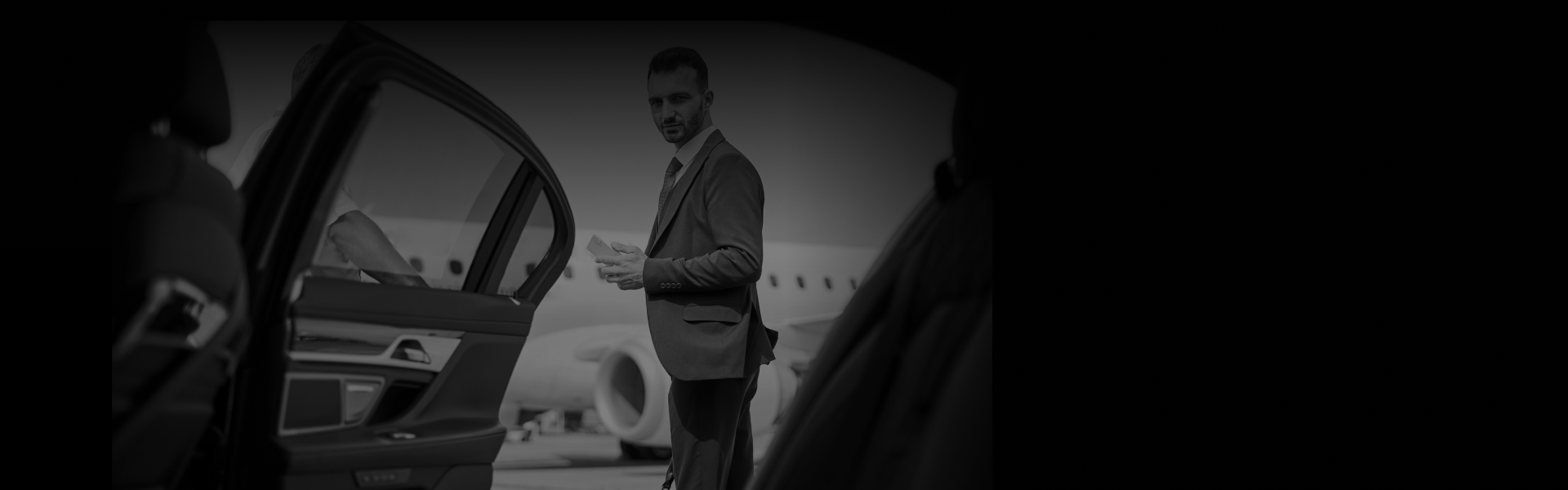 Business traveler in a grey suit exits a luxury black car on the airport tarmac, highlighting premium airport car service from a side rear-door angle.