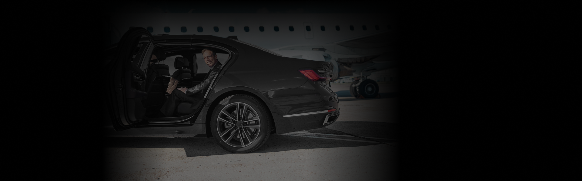 Man in a suit seated in a sleek black car near an airplane, captured from a side-rear angle to highlight the premium Corporate Airport Transfers experience.